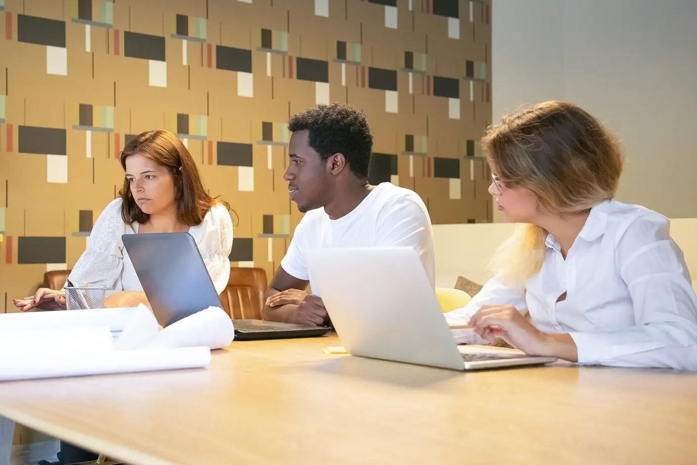 Разнообразная группа дизайнеров сидит за столом с ноутбуками и чертежами, вместе работает над проектом и разговаривает.